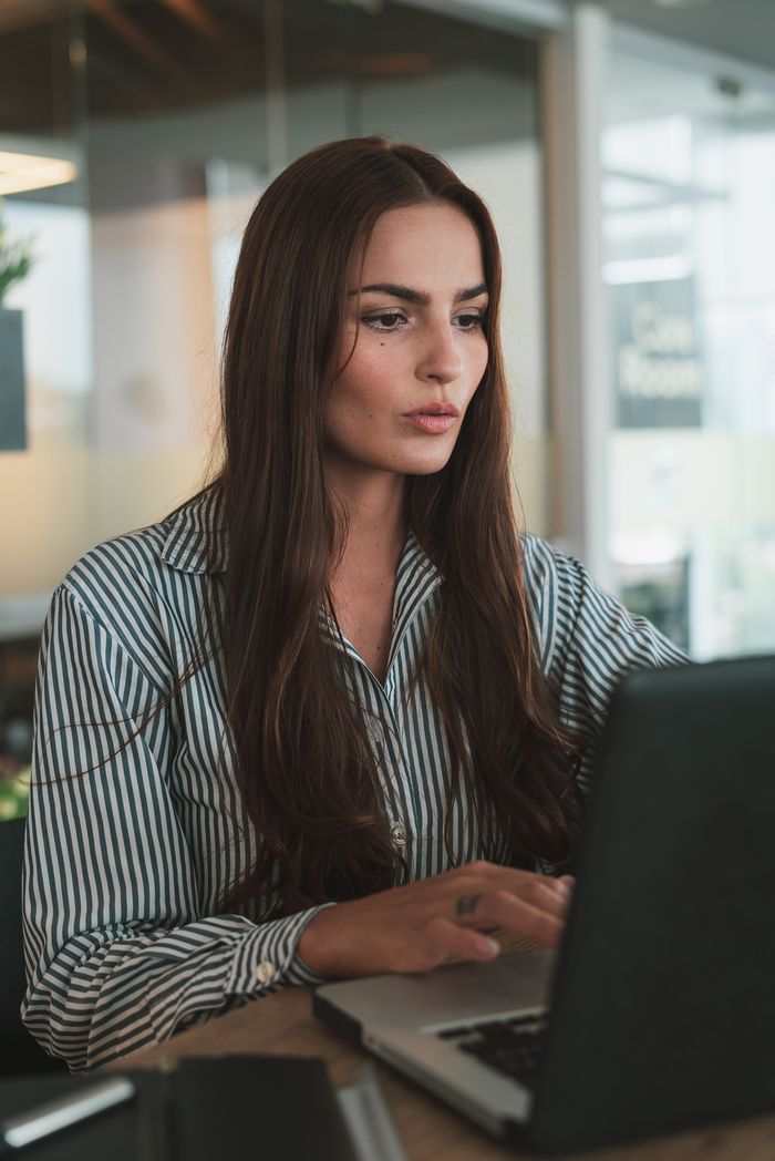 Focused businesswoman with long hair working on a laptop in a modern office environment.